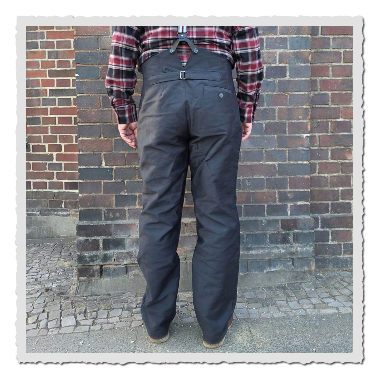 Rückansicht der Arbeiterhose Gustav I mit Cinch-Back und Gesäßtaschen in dunkelgrauem Stoff vor Ziegelwand.